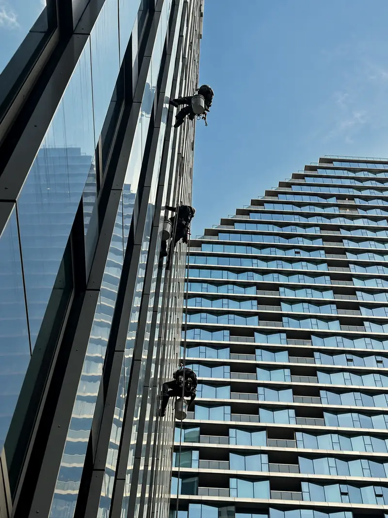 Three rope-access technicians suspended beside a glass facade and a neighbouring tower